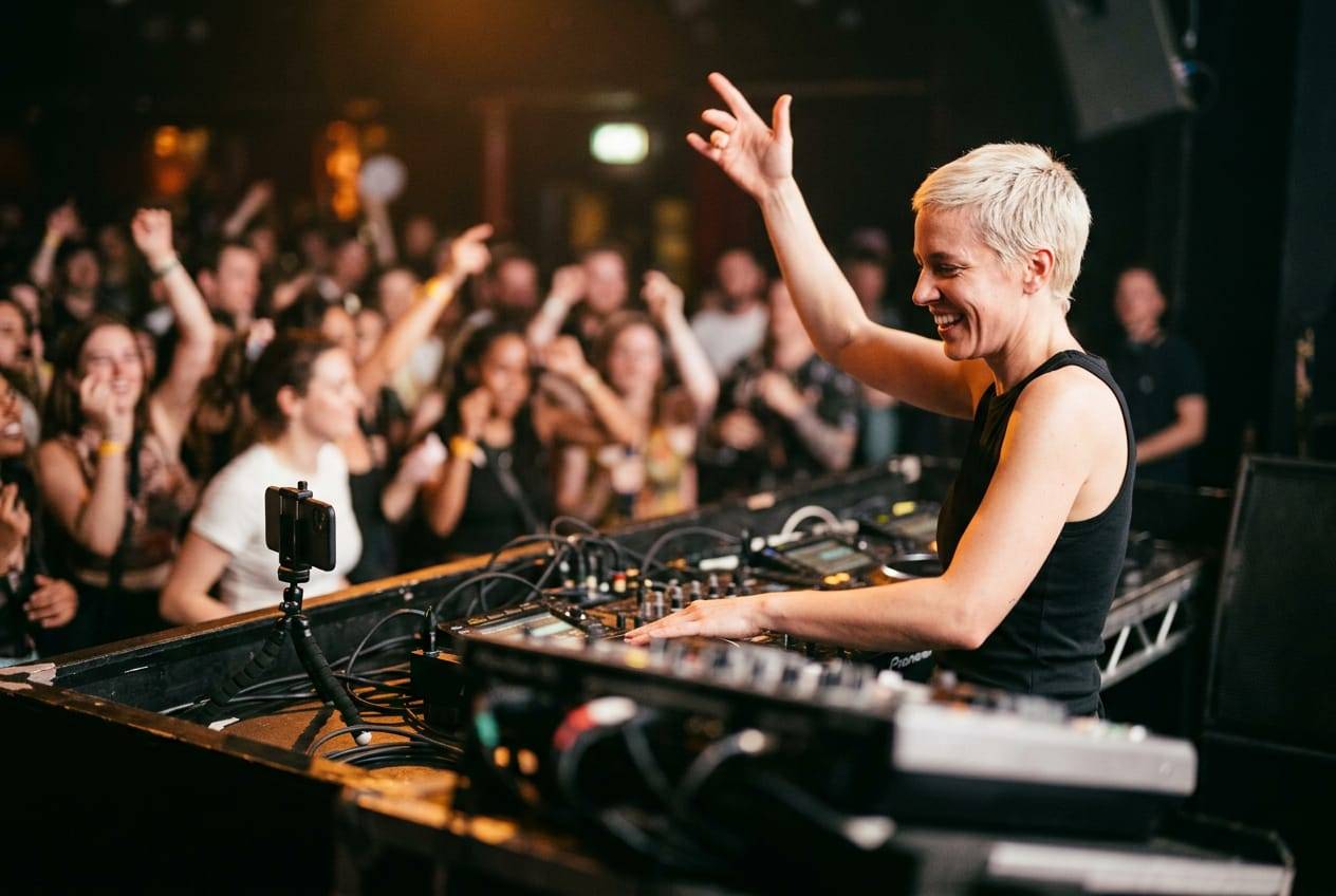 DJ with bleached hair raising a hand to a cheering crowd while a phone on a stand streams her set